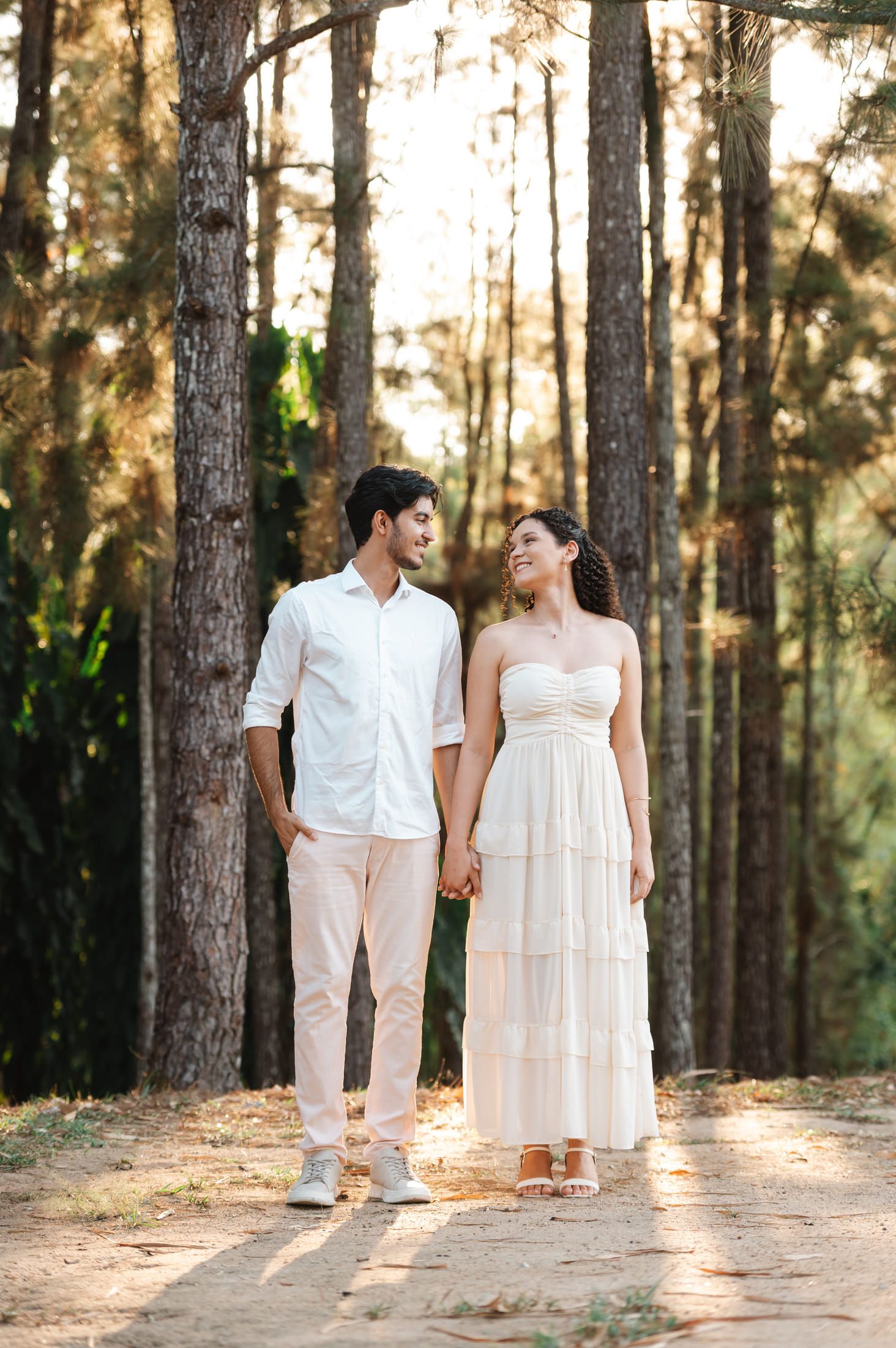 Maria e Italo de mãos dadas na trilha do bosque, sorrindo um para o outro